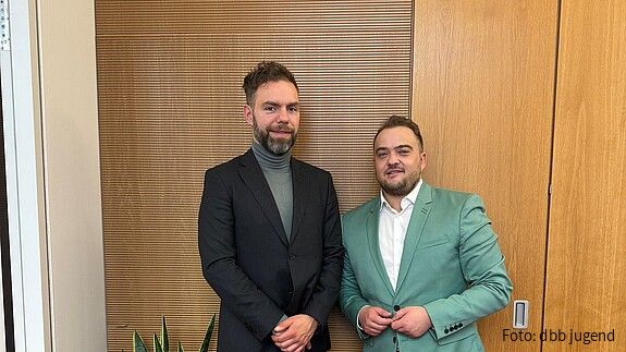 Zwei Männer in Anzügen stehen vor einer Holzwand und blicken in die Kamera